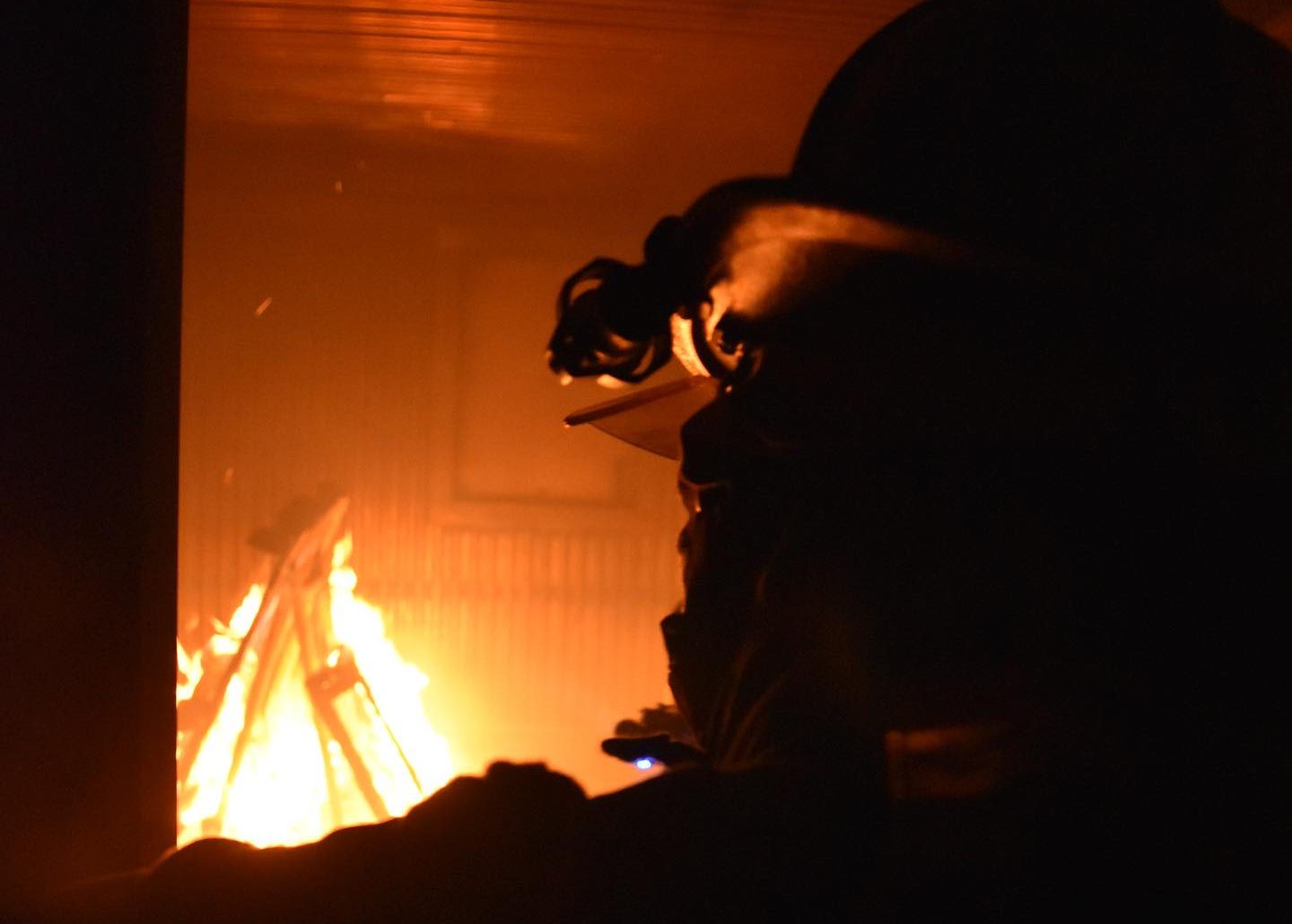 Silhouette of a firefighter wearing a helmet, standing near a large indoor fire. The flames illuminate the background, highlighting the firefighter’s profile and part of the gear. Shadows and smoke create a dramatic scene.