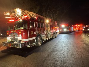 A row of fire trucks with flashing lights is parked on a dark road during nighttime. The trucks are red with white details and have "MIDWAY" written on them. Trees line the road in the background.