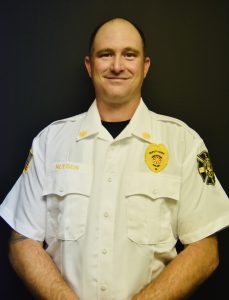 A person in a white uniform shirt stands against a dark background. The shirt has patches and the name "Reeser" on it. The person is smiling and has a short haircut.