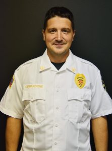 A person in a white uniform with badges and a name tag stands against a dark background. They are looking forward with a neutral expression.