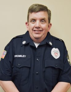 Uniformed individual smiling, wearing a dark shirt with a badge and name tag. The shirt features embroidered patches, including an American flag on the sleeve. The background is plain and neutral.