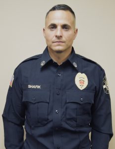 A uniformed man stands against a plain background. He is dressed in a dark uniform with badges and a name tag reading "Shafik." His shirt displays an American flag patch and an emblem with the text "Captain.