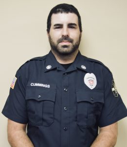 A man in a dark uniform stands against a plain background. His uniform includes patches and a name badge. He appears to be a member of a public service organization.
