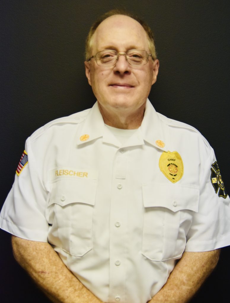 A person wearing a white uniform with badges and patches stands against a dark background. The uniform has "Chief" and "Fleischer" on it. The person is smiling slightly, and their arms are crossed.
