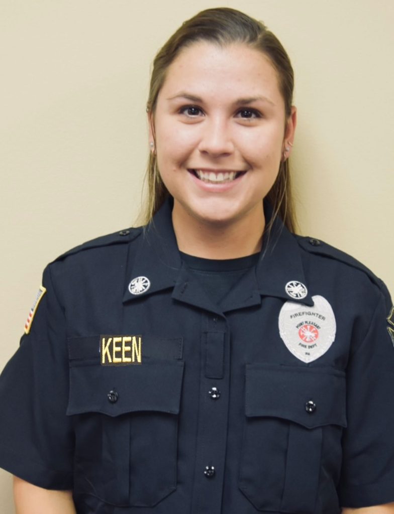 A person in a black uniform with a name patch smiles against a beige background. The uniform features insignia and a patch indicating they are a firefighter.