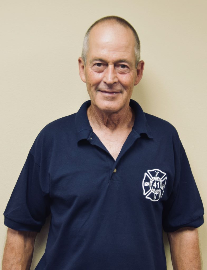 An older adult wearing a navy blue polo shirt with a fire department logo and the number 41 stands against a light beige wall, smiling slightly.
