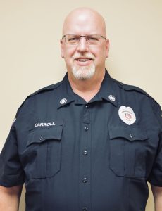A man with glasses and a goatee wearing a black uniform with a badge and the name "Carroll" embroidered on it stands in front of a plain light-colored background, smiling.