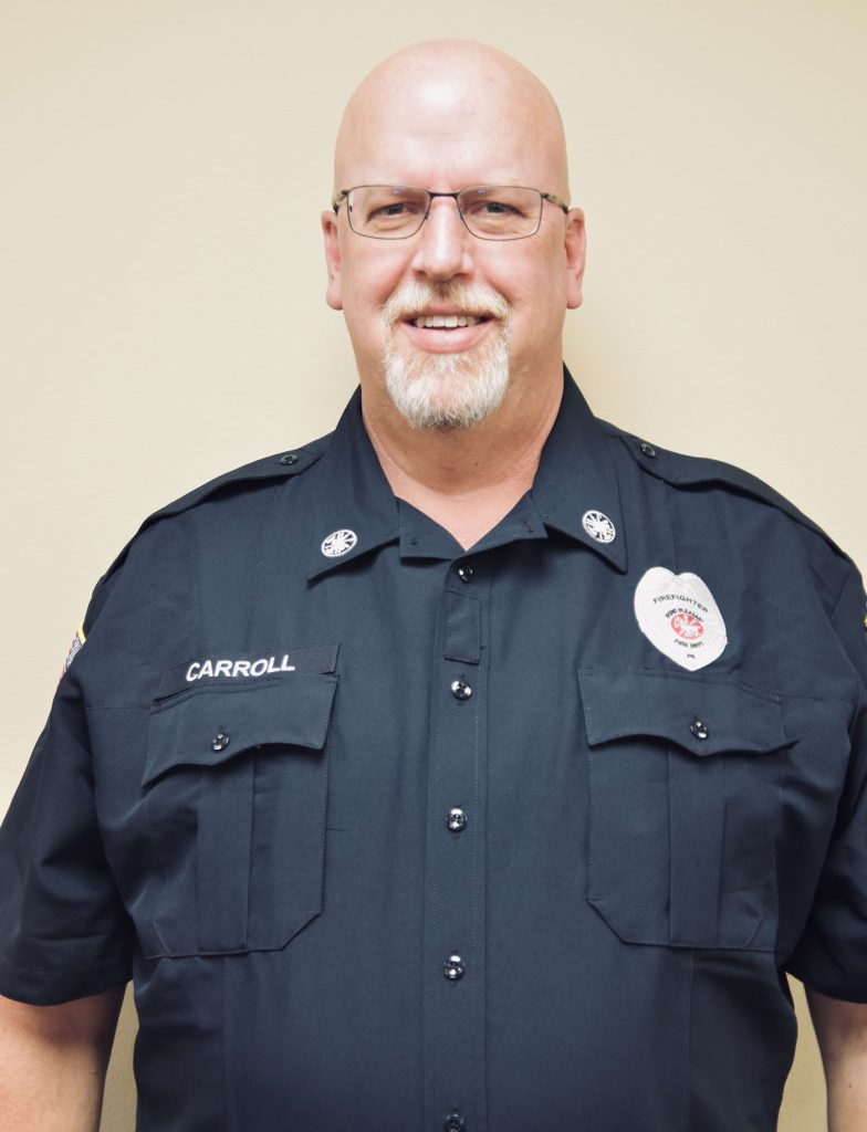 A man with glasses and a goatee wearing a black uniform with a badge and the name "Carroll" embroidered on it stands in front of a plain light-colored background, smiling.
