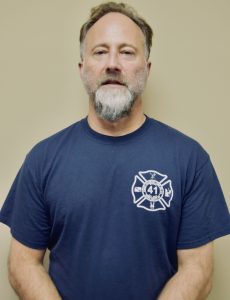 A man with short hair and a beard is standing against a plain background. He is wearing a navy blue T-shirt with a firefighting emblem on the chest, displaying the number 41.