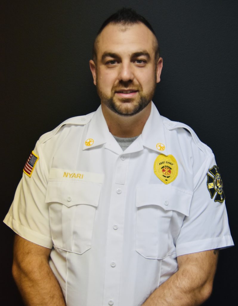 A person in a white uniform with badges stands against a black background. The uniform features an American flag patch, a name tag reading "Nyari," and two fire department emblem patches. The individual has short dark hair and a beard.