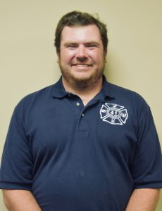 A person with short hair and a beard is smiling, wearing a navy blue polo shirt with a white emblem on the chest, standing against a beige background.