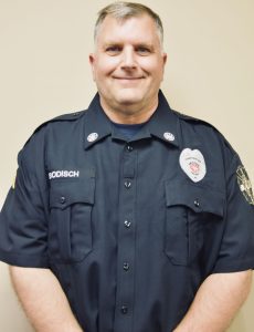 A person in a navy blue uniform with badges and patches stands against a light-colored wall, smiling.