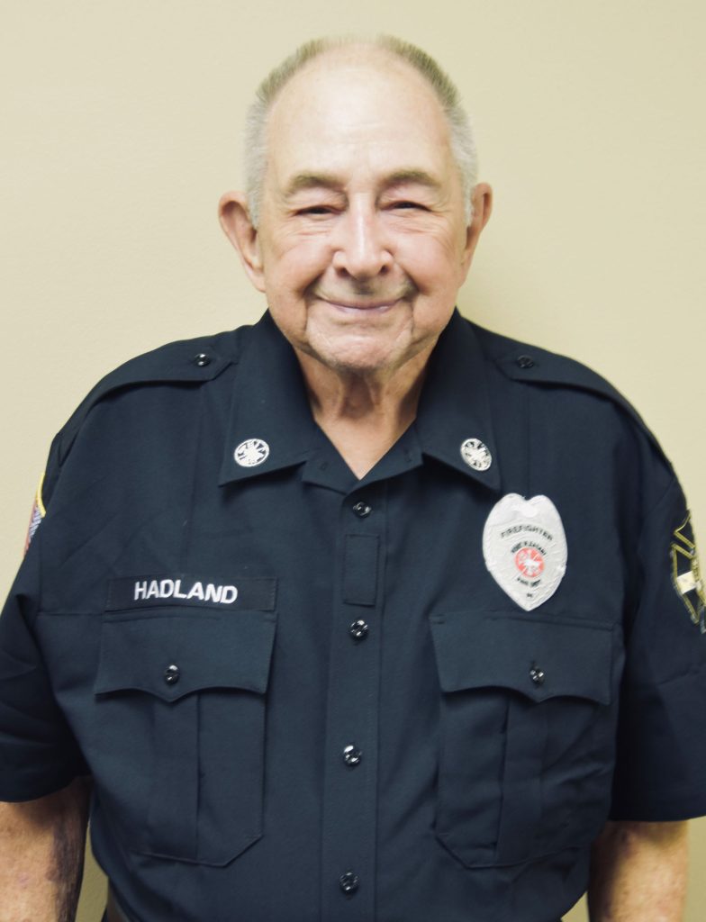 An elderly man with short gray hair smiles while wearing a dark uniform with badges. His name tag reads "Hadland." He stands against a plain, light-colored background.