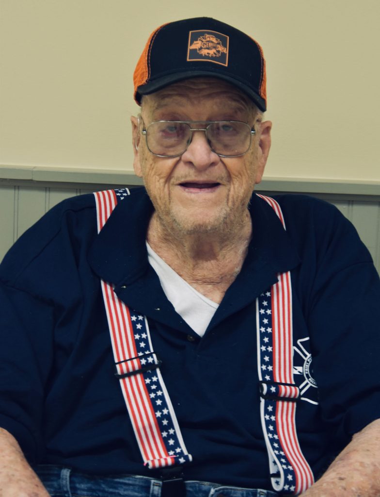Elderly man smiling, wearing a black cap and glasses. He's dressed in a navy shirt with a logo and star-spangled suspenders. Background shows a beige wall with vertical paneling.