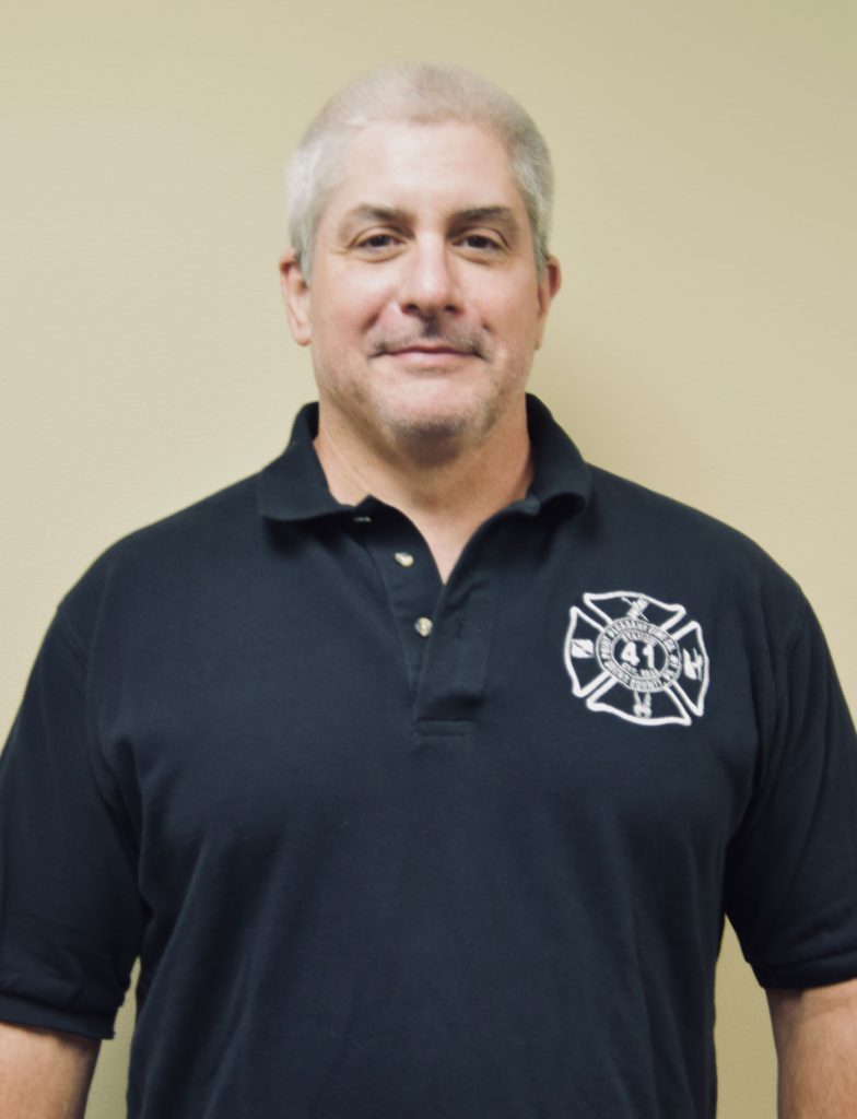 Man with short gray hair wearing a black polo shirt with a fire department emblem, standing against a plain beige background.