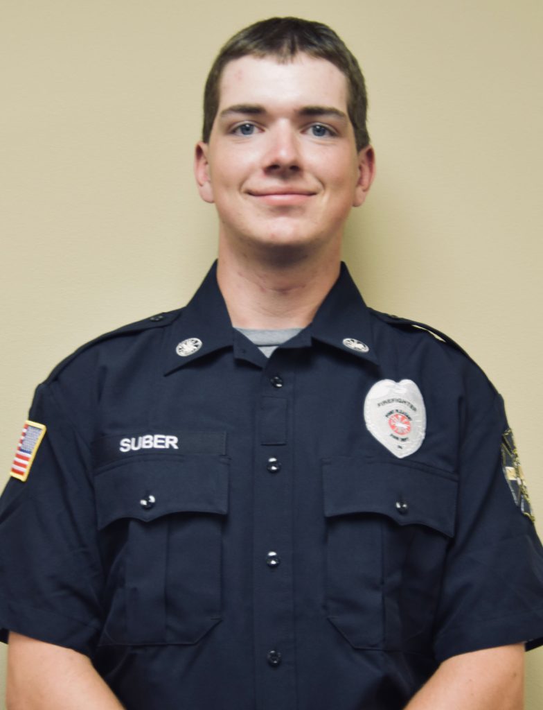 A person wearing a police uniform stands against a plain background. The uniform has a badge, shoulder patches, and name tag. The person has short hair and a slight smile, with arms relaxed by their sides.