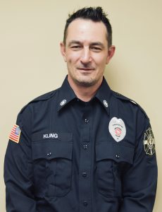 A person wearing a dark uniform with fire department insignia and the name "Kling" on the chest, standing against a plain beige background. They have short dark hair and a neutral expression.