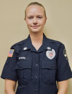 A person in a dark police uniform stands against a neutral background. The uniform features badges, patches, and an American flag on the sleeve. The person has blonde hair and is looking at the camera with a neutral expression.