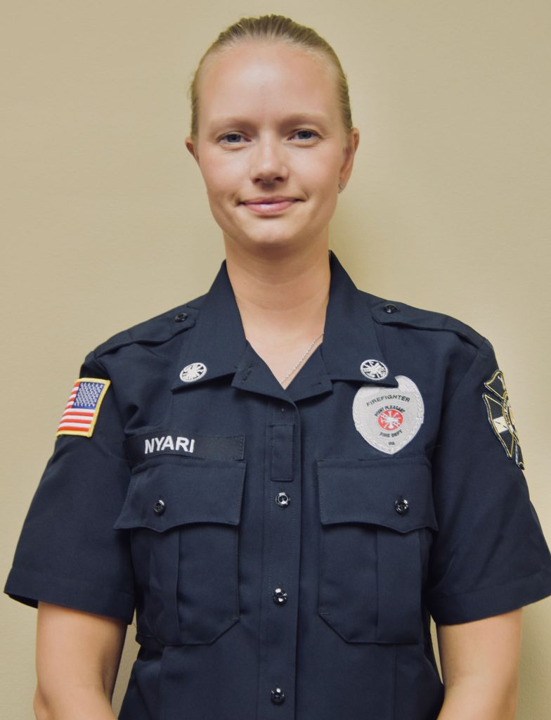 A person in a dark police uniform stands against a neutral background. The uniform features badges, patches, and an American flag on the sleeve. The person has blonde hair and is looking at the camera with a neutral expression.