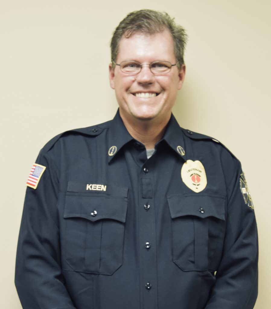 A smiling person wearing glasses and a dark uniform with badges stands against a beige wall. The uniform features a name tag with "KEEN" and a patch with the U.S. flag.