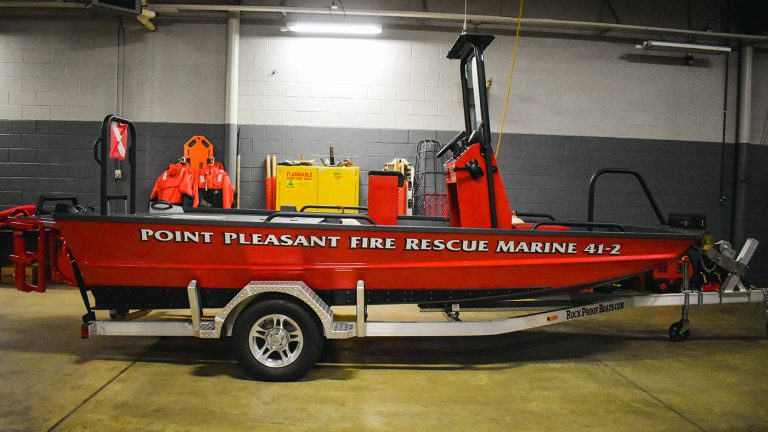 Red rescue boat labeled "Point Pleasant Fire Rescue Marine 41-2" on a trailer indoors. The boat has a steering console, emergency lights, and equipment storage; a gasoline container and flotation devices are in the background.