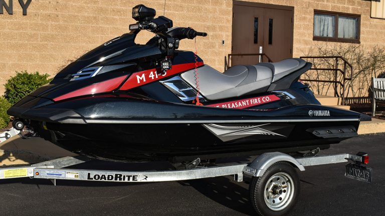 A black and red Yamaha jet ski is mounted on a Load Rite trailer outside a building. The jet ski is marked with "M 41-1" and features a camera on top. It is labeled with "Pleasant Fire Co." and parked on a paved area.
