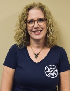 A person with curly blonde hair and glasses smiles at the camera. They are wearing a navy blue t-shirt with a "41" emblem, featuring a fireman's helmet and crossed axes, on the chest. The background is a plain beige wall.