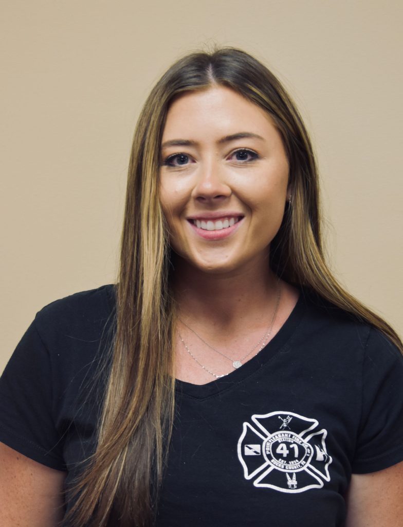 A person with long brown hair is smiling, wearing a black shirt with a firefighting emblem featuring the number 41. The background is a plain beige wall.