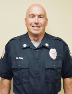 A man in a navy blue uniform stands against a neutral background. The uniform has a badge and patches. The man has a friendly expression and short, light-colored hair.