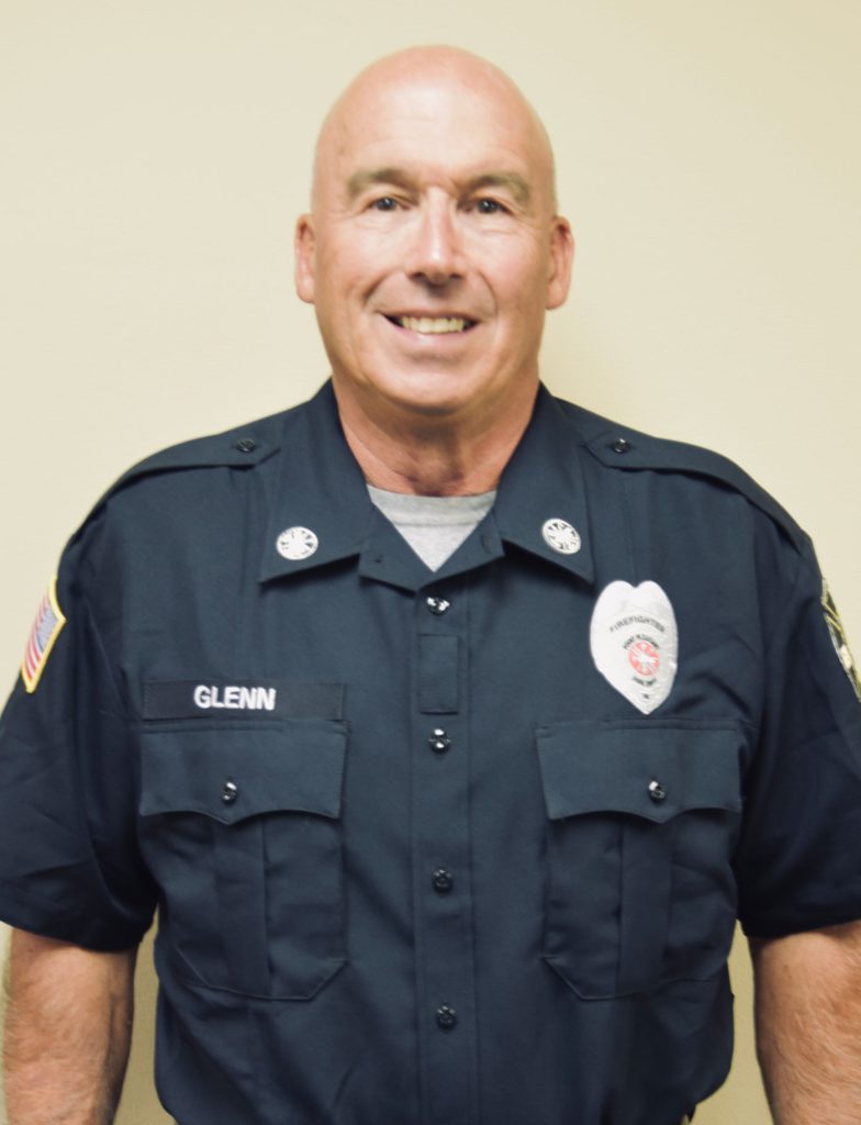 A man in a navy blue uniform stands against a neutral background. The uniform has a badge and patches. The man has a friendly expression and short, light-colored hair.