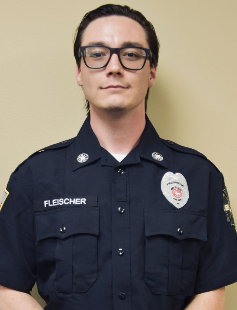 A person in a dark police-style uniform stands against a neutral background. The shirt displays a badge and the name "Fleischer." They have short, dark hair and are wearing glasses.