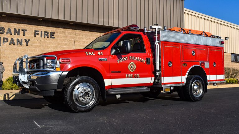 A red fire truck labeled "Point Pleasant Fire Co." is parked on a sunny day outside a beige brick fire station. The truck is equipped with emergency lights, a hose, and additional firefighting equipment.