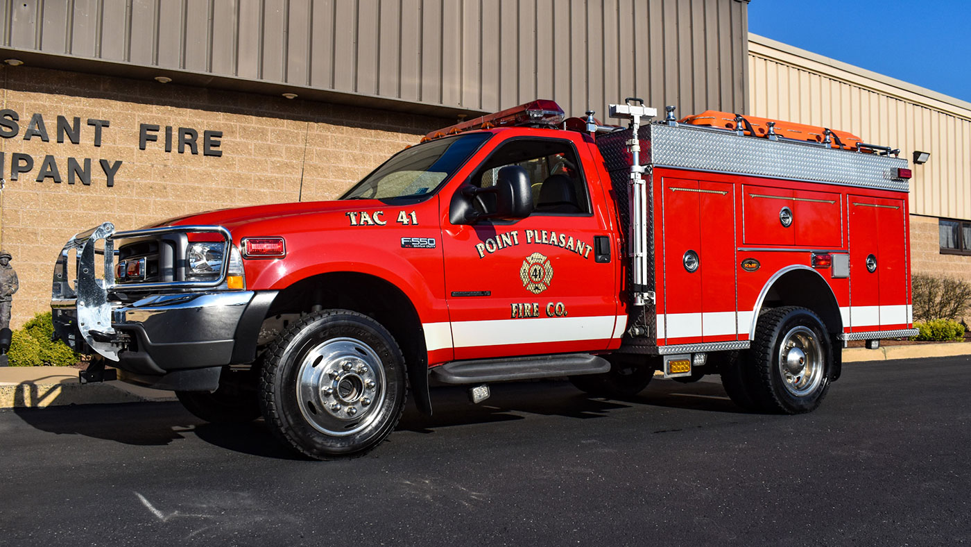 A red fire truck labeled "Point Pleasant Fire Co." is parked on a sunny day outside a beige brick fire station. The truck is equipped with emergency lights, a hose, and additional firefighting equipment.