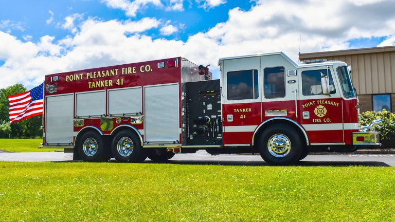 A bright red fire truck labeled "Point Pleasant Fire Co., Tanker 41" is parked on a grassy field. An American flag is attached to the back. The sky is blue with scattered clouds.