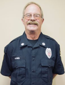 A smiling older man with glasses and a mustache is wearing a dark blue uniform with badges and a name tag that reads "KLING." The uniform includes a badge indicating he is part of a fire department. The background is a plain beige wall.