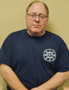 A man stands against a light brown wall wearing glasses and a navy blue t-shirt with a white emblem featuring the number 41. His arms are resting in front of him casually.