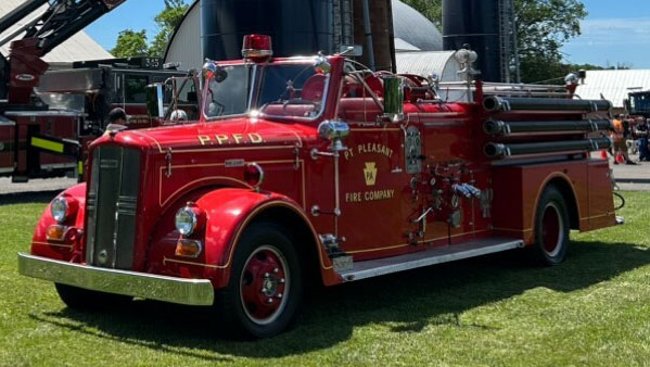 A vintage red fire engine with "P.P.F.D." and "Mt. Pleasant Fire Company" on the side, parked on grass. The truck has hoses and ladders, with a shiny exterior and a classic design. Background includes other fire trucks and trees.