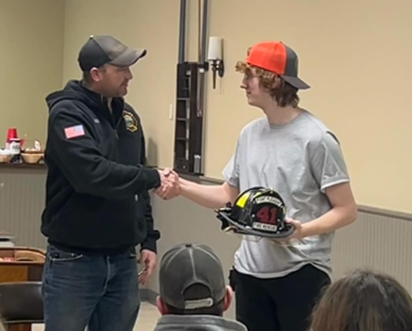 Two men shaking hands indoors. One man, wearing a black hoodie and cap, has an American flag patch on his sleeve. The other, in a gray shirt and backward cap, holds a firefighter helmet with "41" on it. Onlookers are seated in the foreground.