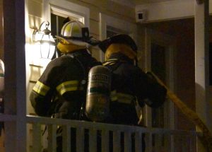 Two firefighters in gear and helmets stand on a porch at night, lit by an outdoor lamp. One carries a hose, ready to respond to an emergency. The house has light-colored siding and a decorative railing.
