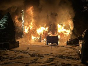 A large house engulfed in flames at night, with thick smoke rising into the sky. A pickup truck is parked in the snowy driveway. The scene is illuminated by the intense fire, surrounded by snowy ground and trees.