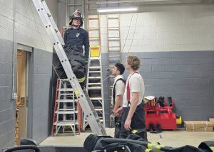Three firefighters are in a garage-like room with one on a tall ladder against a wall. Two others stand nearby watching. Firefighting gear is scattered on a table in the foreground. The setting appears to be a fire station with equipment around.