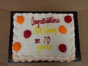 A rectangular cake with white frosting and orange and red decorations. The text reads, "Congratulations Kid & Janey on 70 years" in red and yellow icing. The cake is placed inside a cardboard box.