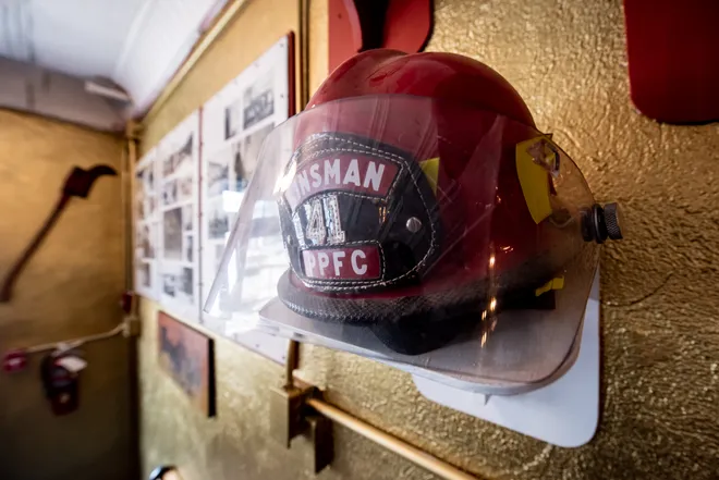 A firefighter's helmet with a clear visor and labeled "FIREMAN 141 PPFC" is mounted on a gold-textured wall. Nearby, there are framed photographs and firefighting tools displayed.
